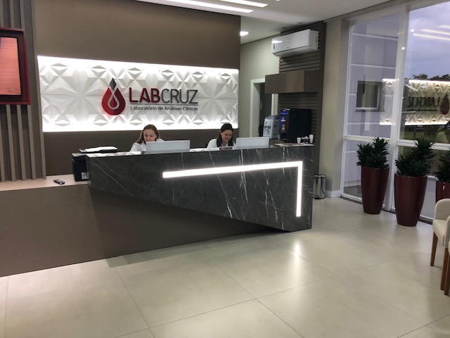 Foto da parte interna da entrada do laboratório LabCruz, duas atendentes estão sentadas atrás do balcão de atendimento baixo do logotipo do laboratório, que está fixado na parede.